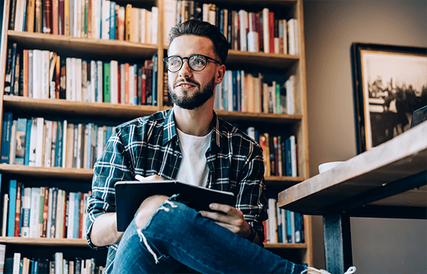 Contemplative male author in optical eyeglasses holding notepad for writing in hands and looking off to the side inside of a library