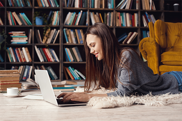 A woman laying down on the ground working on a laptop inside of an at home library. There is a large bookshelf and half of a yellow couch in the background.